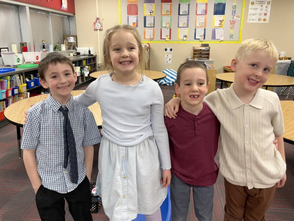 Four young students stand with their arms around each other's shoulders, smiling for a camera in a classroom. They are dressed in light-colored shirts and dresses.