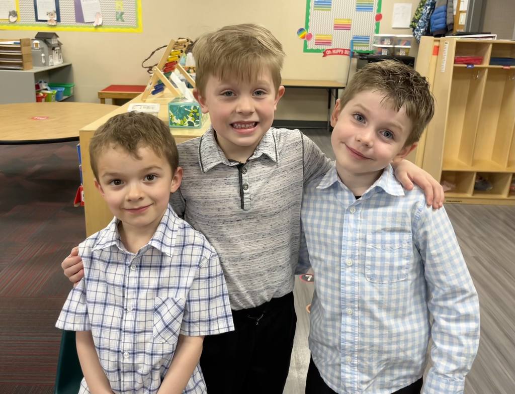 Three young boys pose together with their arms linked in a classroom, all smiling widely. They are wearing button-up shirts, two in plaid or checkered patterns.