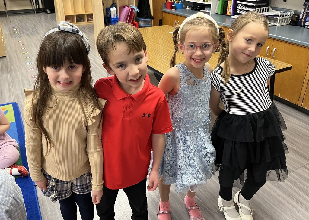 Four students—three girls in nice dresses and one boy in a red polo shirt—pose closely together in a classroom, all smiling broadly.