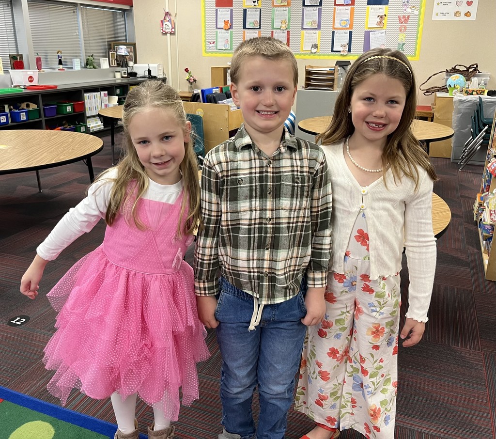 Three smiling young children pose for a picture in a classroom. A girl in a pink ruffled dress holds hands with a boy in a plaid shirt.