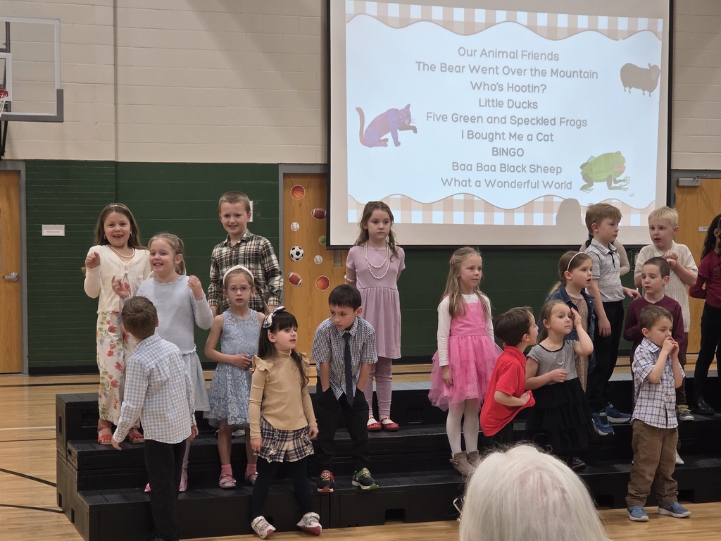 A group of young students stands on risers in a gymnasium for a performance, wearing nice outfits. A large projector screen behind them lists a song program with cartoon animals.