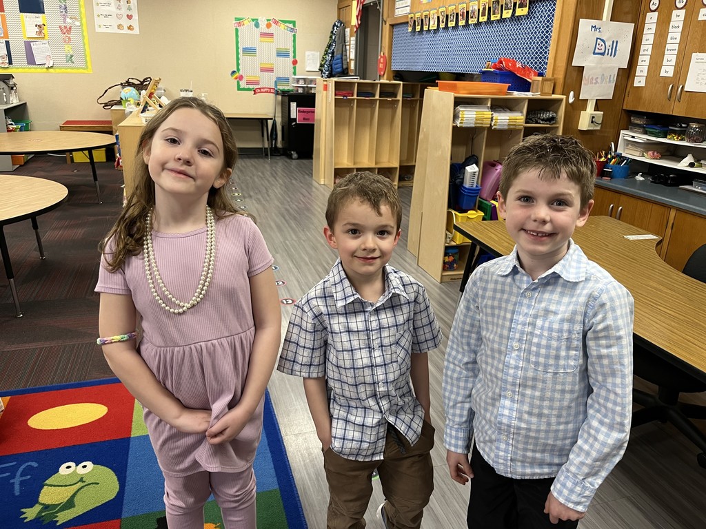 Three smiling students, one girl and two boys, pose together for a photo in a classroom. The girl wears a purple dress and a double-strand pearl necklace.