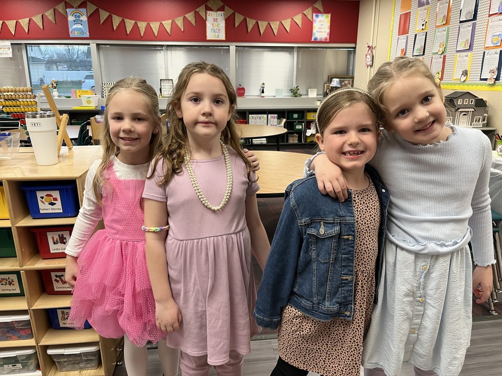 Four young girls stand together in a classroom, showing a close bond by leaning against each other. They wear brightly colored and patterned dresses and shirts.