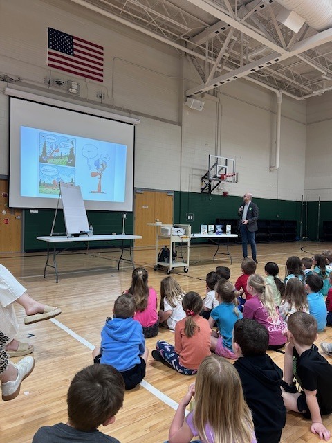 An author stands in a school gymnasium giving a presentation to a large group of elementary students seated on the floor. A projector screen displays a comic strip, and several children's books are lined up on a table.