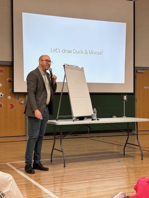 A man in a blazer and glasses smiles while holding a microphone and standing next to a white drawing easel. The projector screen behind him reads, "Let's draw Duck & Moose!"