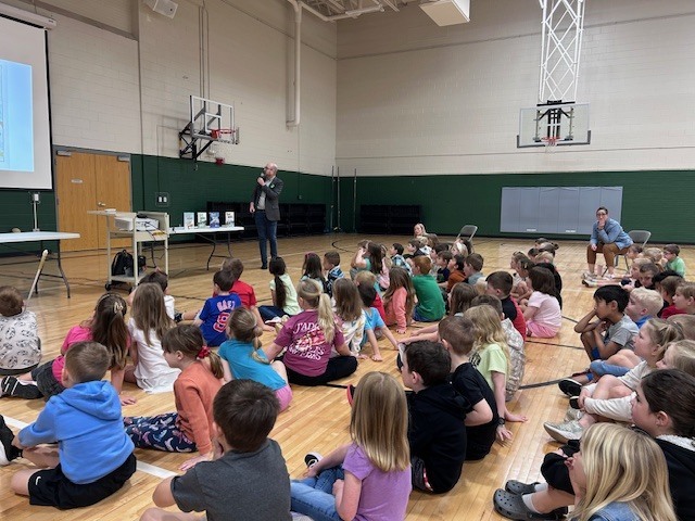 A wide shot of a school gymnasium showing dozens of children sitting on the polished wood floor, looking toward a presenter and a projection screen during an assembly.