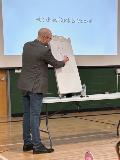A man uses a blue marker to draw a character on a large paper easel during a presentation. The background screen displays the text "Let's draw Duck & Moose!"
