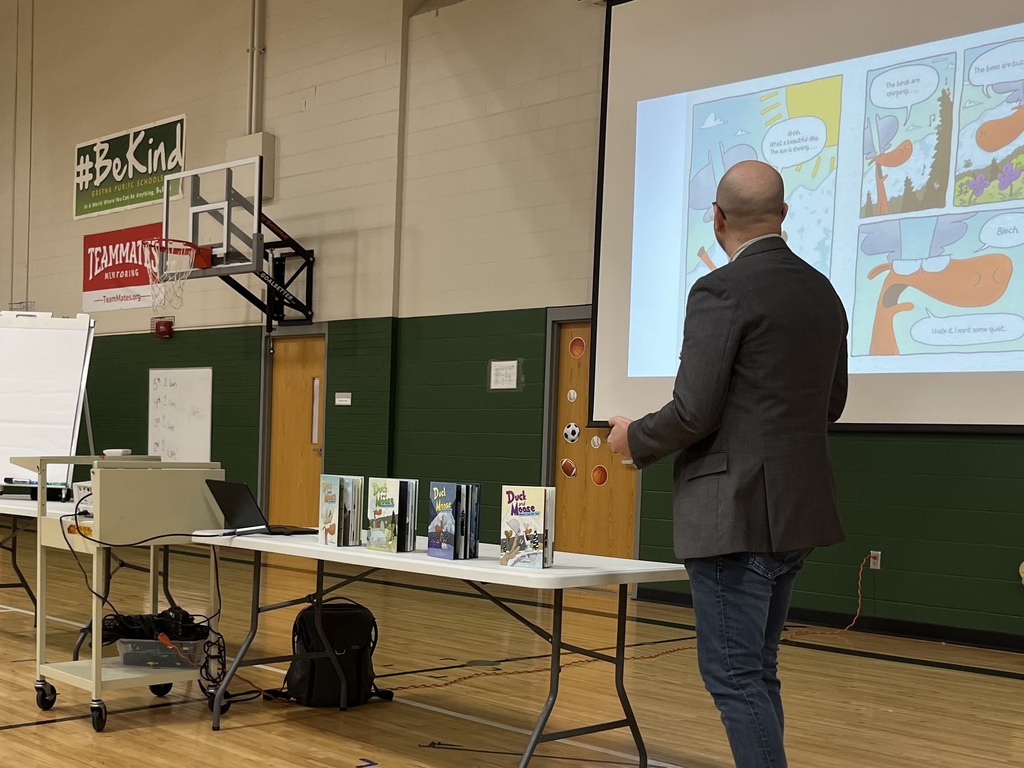 A man stands before a projector screen displaying a comic strip about a duck and a moose; several Duck and Moose books are lined up on a table in the foreground.