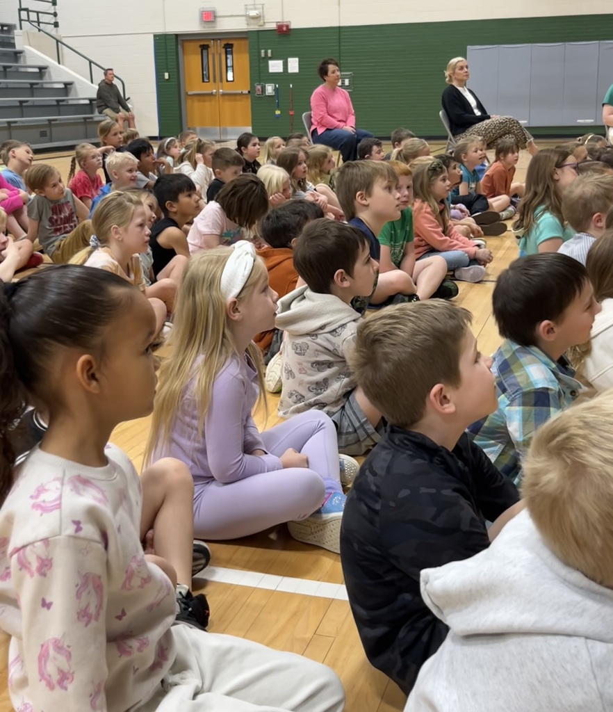A close-up side view of children sitting cross-legged on a wooden gym floor, focused intently on a speaker.