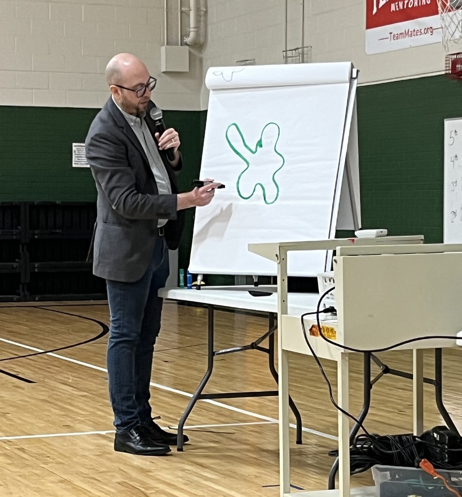 A bald man in a gray blazer and glasses stands in a gym, using a microphone while pointing at a large green doodle on a flip chart.