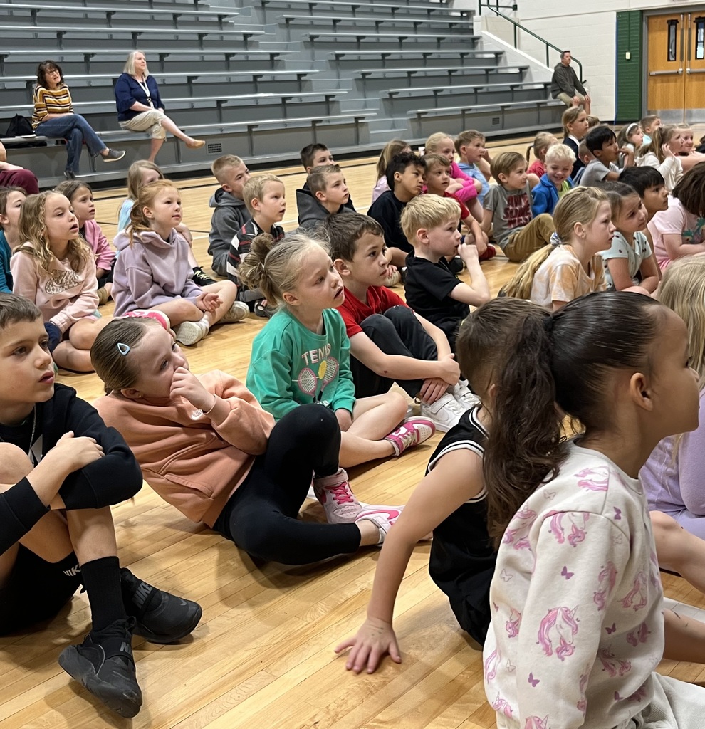 Children sitting on a gym floor looking upward, with adults seated on bleachers in the background.
