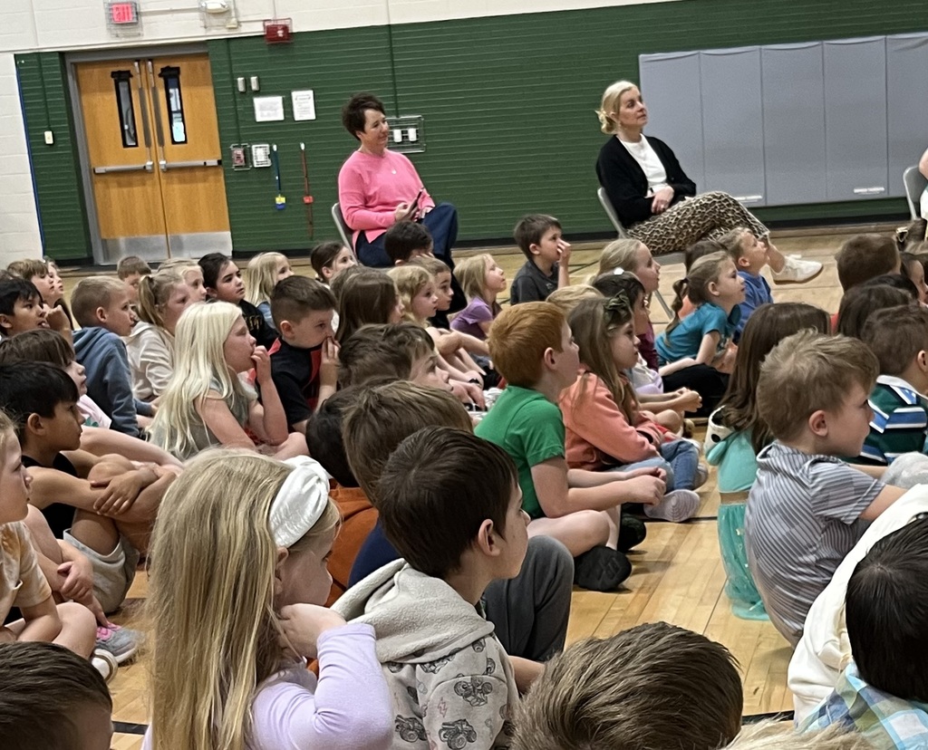 A large group of elementary school children sit on a gymnasium floor, looking toward a presentation off-camera.