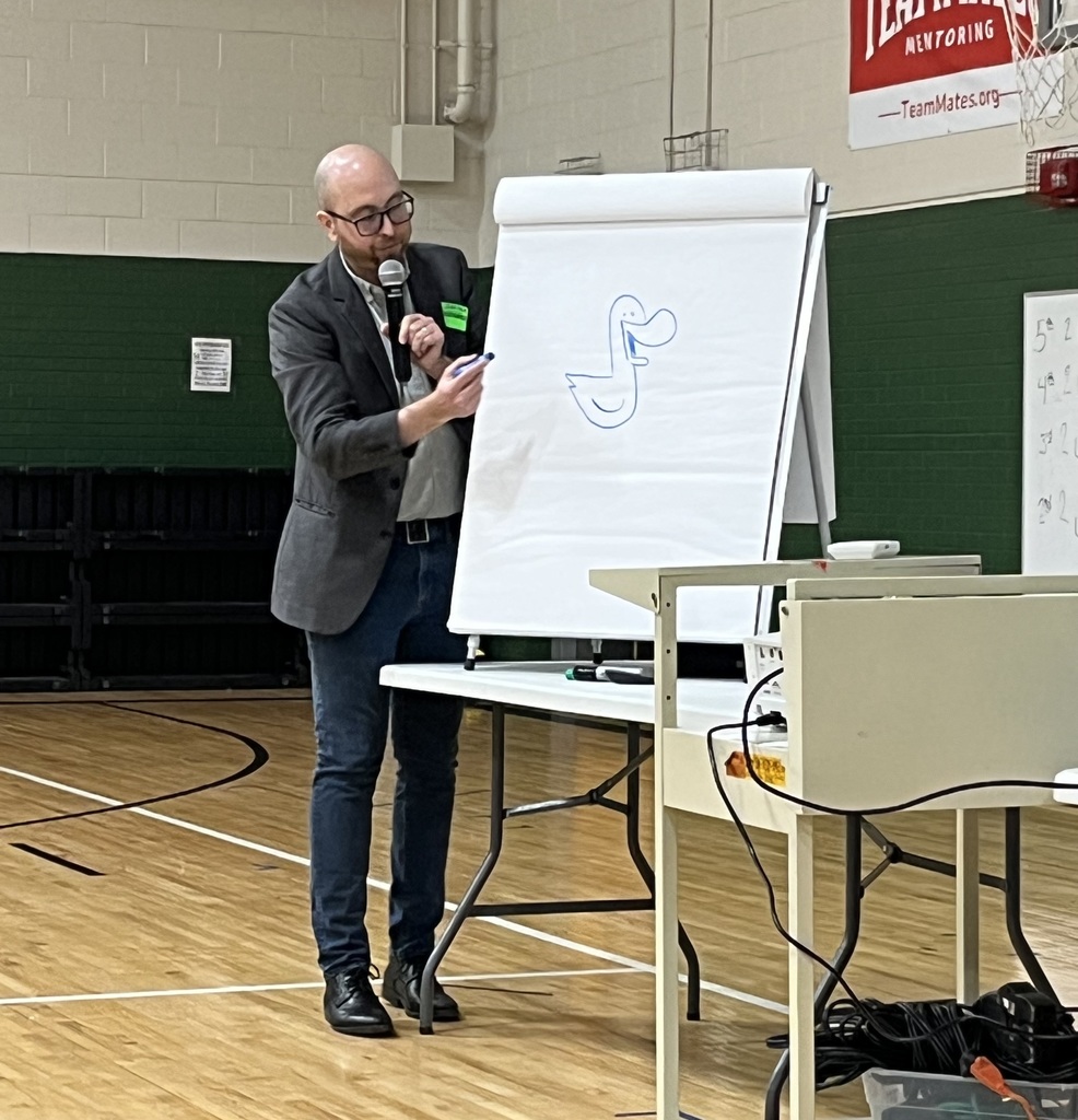A man in a blazer draws a blue cartoon duck on a flip chart while speaking into a microphone in a school gym.