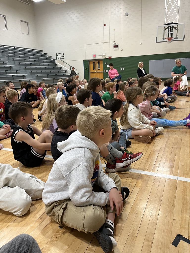 A wide shot of a gymnasium showing rows of children sitting on the floor watching a presentation, with bleachers and a basketball hoop in the background.