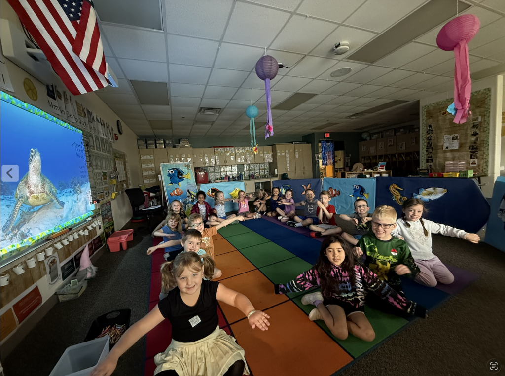 Students are sitting on a rug acting like they are swimming during the ocean habitat rotation.