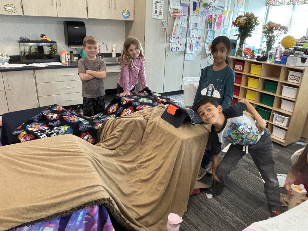 Two girls and two boys are standing next to their blanket fort smiling.