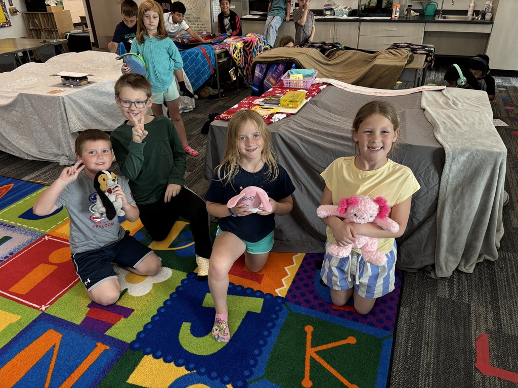 Two girls and two boys kneel next to a blanket fort holding stuffed animals.