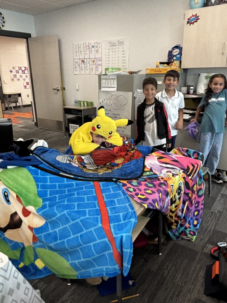 A boy in a pokemon hoodie leans over the top of a blanket fort.  Two boys and a girl stand behind him smiling.