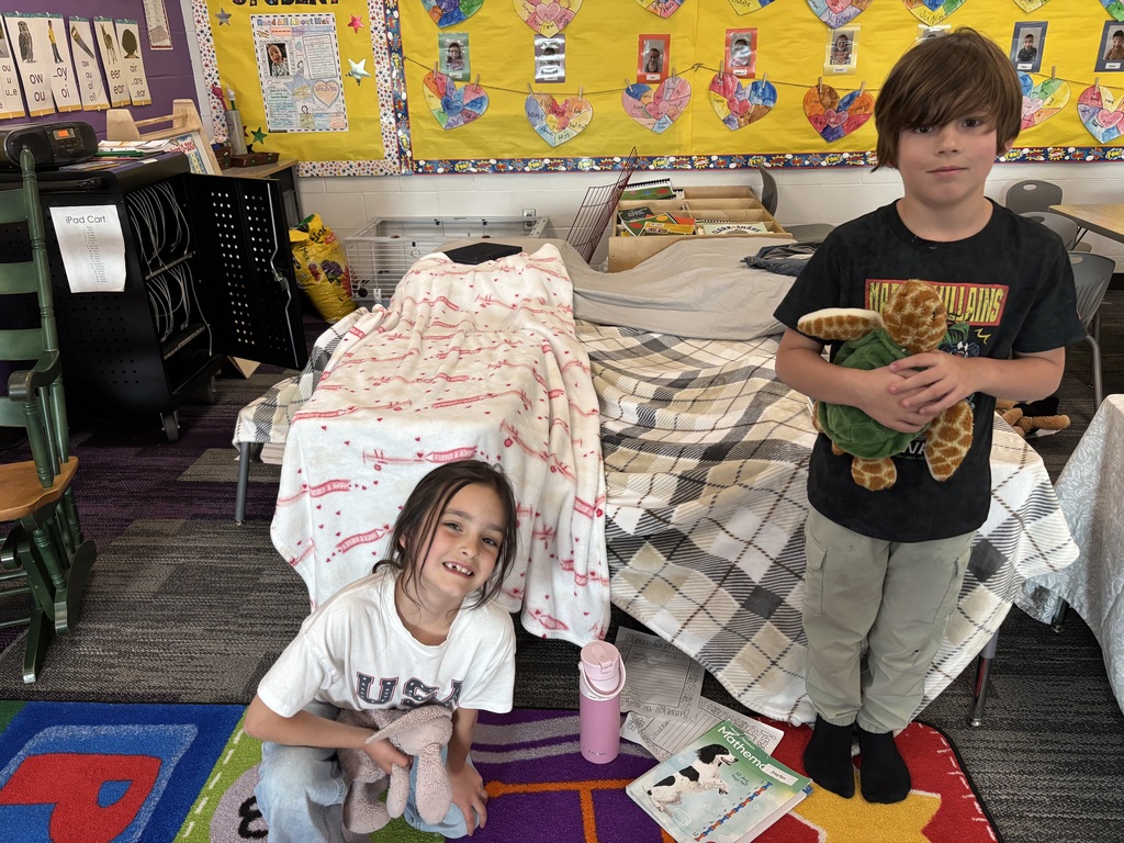 A girl sits in front of a blanket fort and a boy stands next to her.  Both are holding stuffed animals.