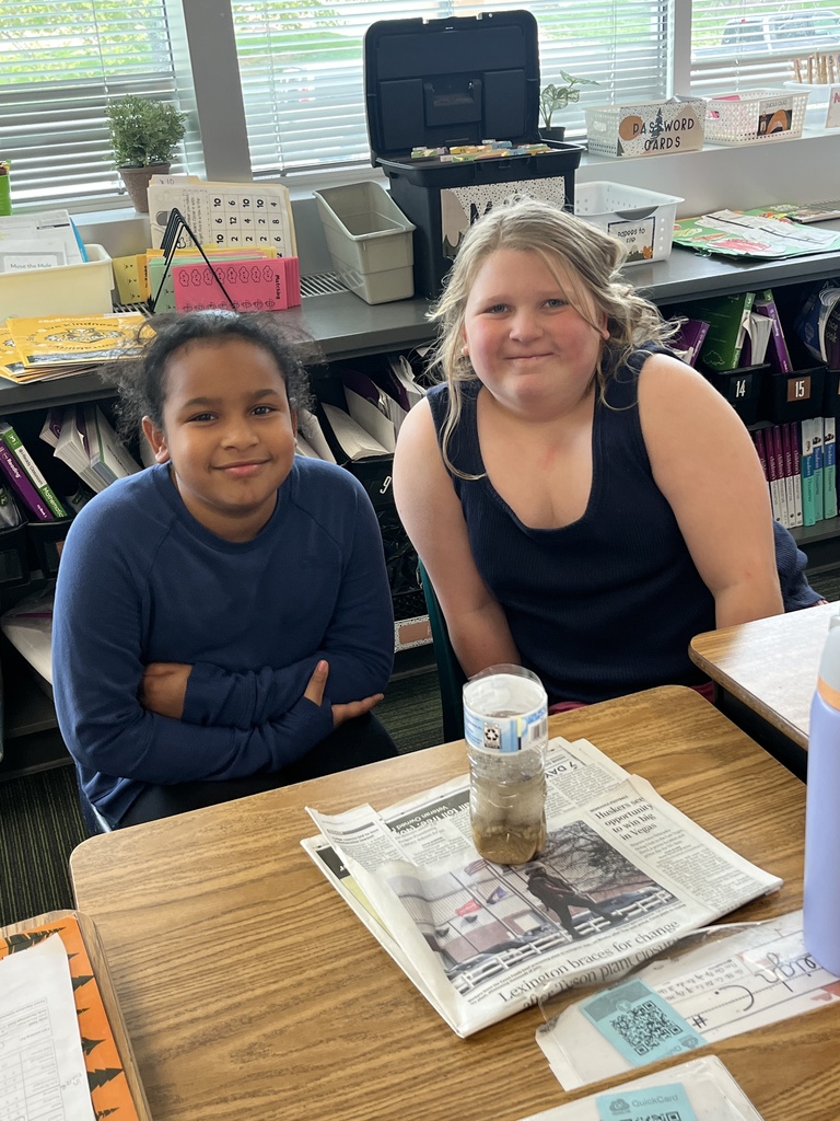 Two girls sit at a desk posing for a photo behind a plastic bottle filtration experiment resting on a newspaper.