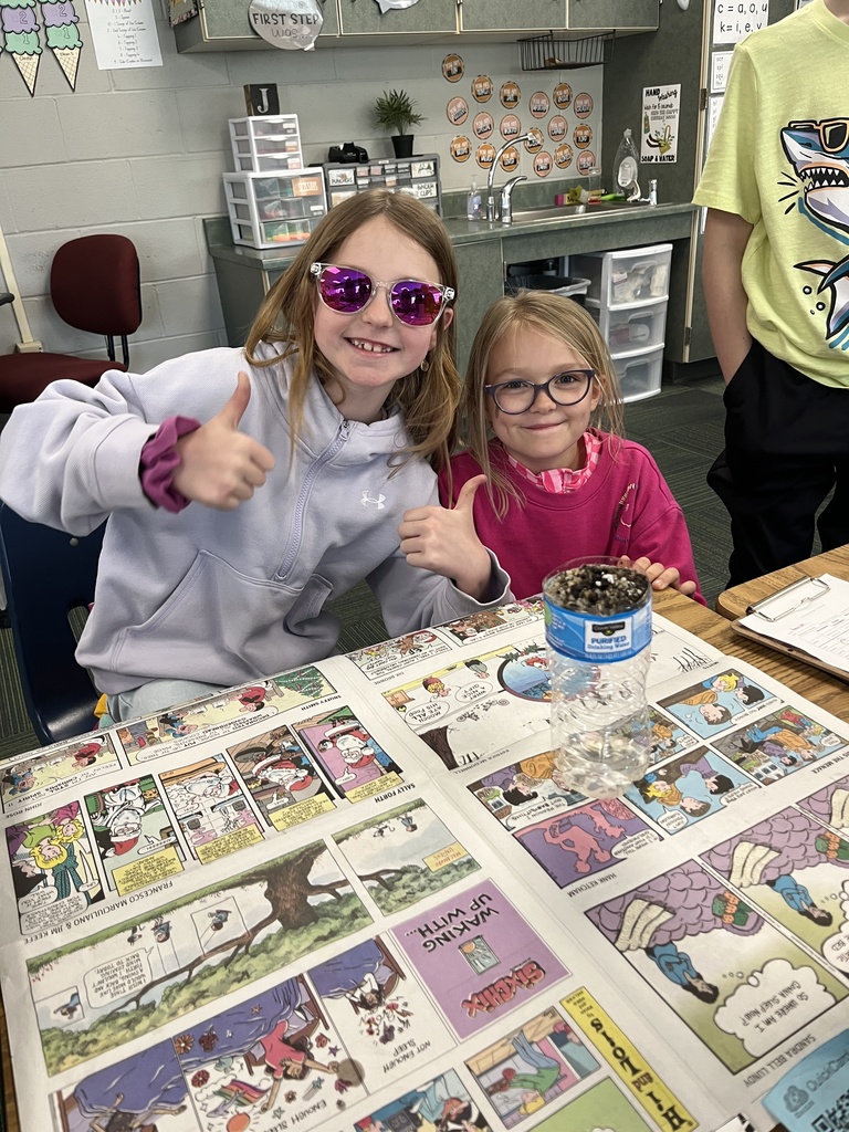 Two young girls give thumbs up behind a desk covered in Sunday comic strips and their filtration project.