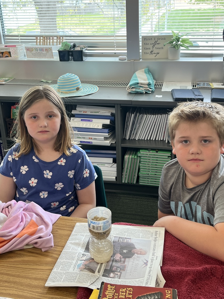 A girl in a floral shirt and a boy in a grey shirt sit behind their completed water bottle filter on a classroom desk.