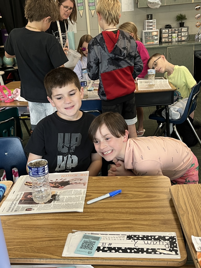 Two young boys in a classroom smile next to a DIY water filtration project made from a plastic bottle. Other students and a teacher work in the background.