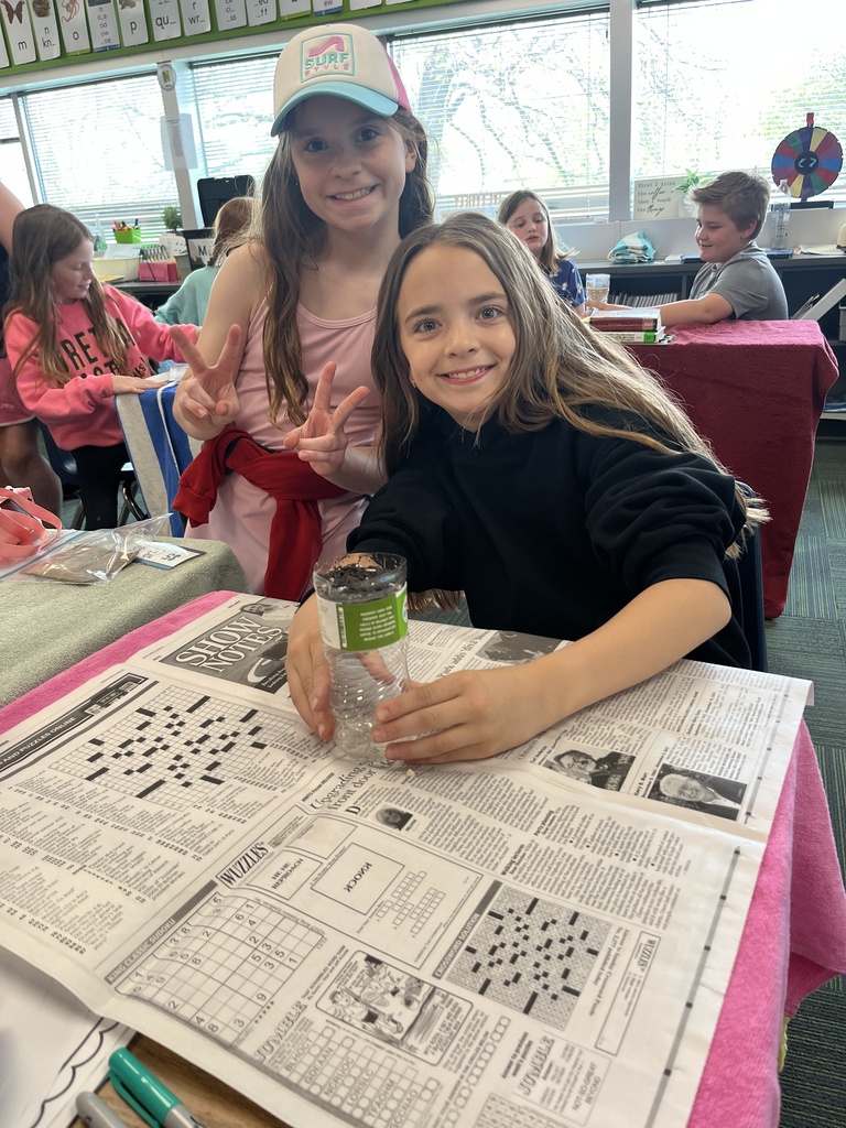Two girls, one wearing a "Surf Style" hat and giving peace signs, pose with their water filter experiment on a desk covered in newspaper puzzles.