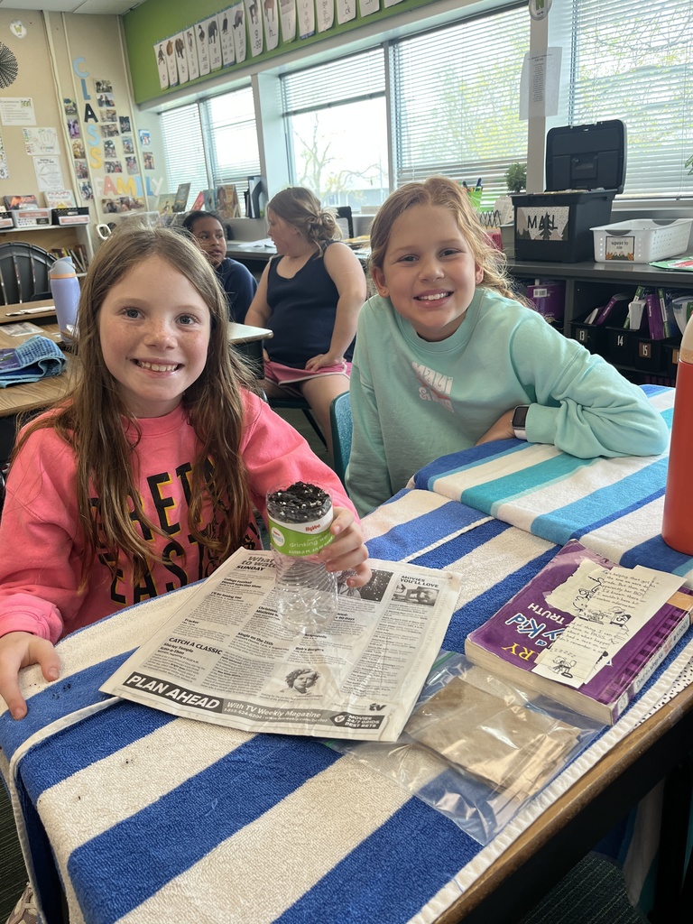 Two smiling girls sit at a desk draped with a blue and white striped towel, displaying their plastic bottle experiment.