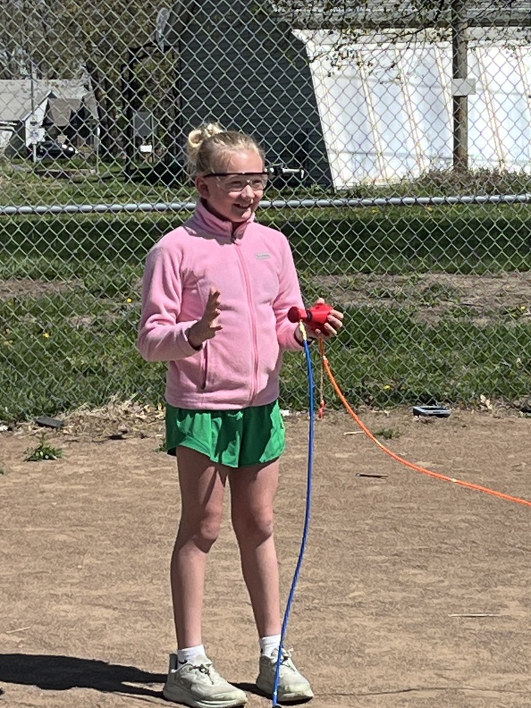 A girl in a pink zip-up and green shorts holds the red rocket launcher while wearing safety eyewear.