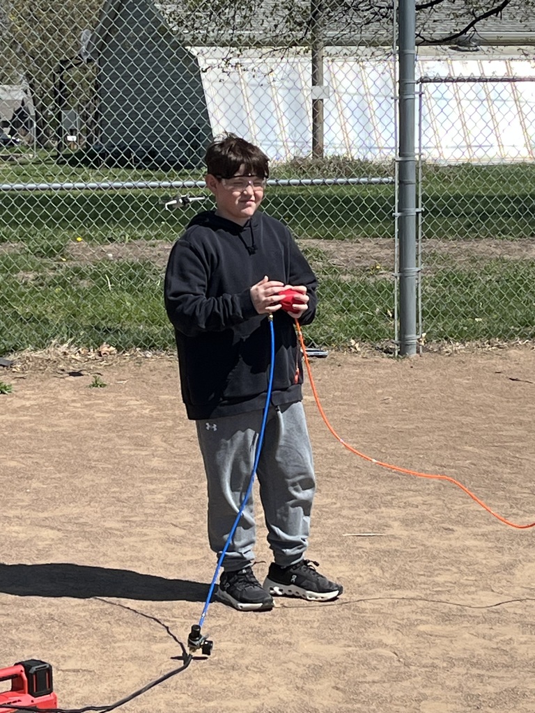 A boy in a black hoodie and safety glasses holds the red rocket launch controller on a baseball diamond.