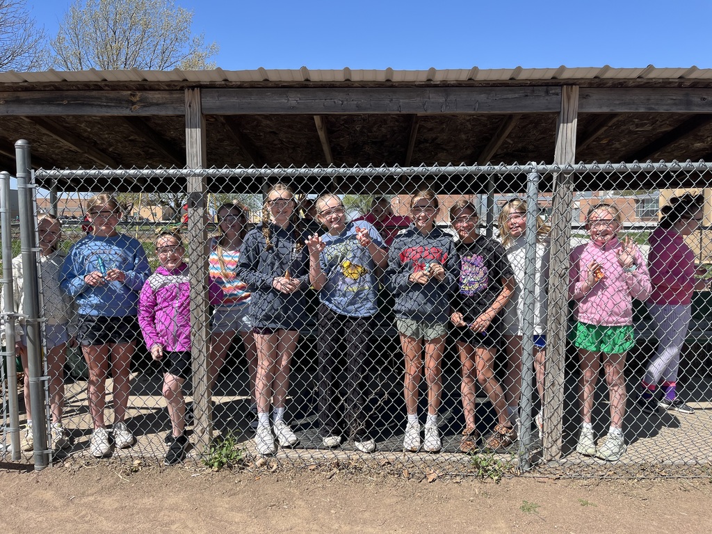 Ten children wearing safety glasses pose for a group photo behind a chain-link dugout fence.