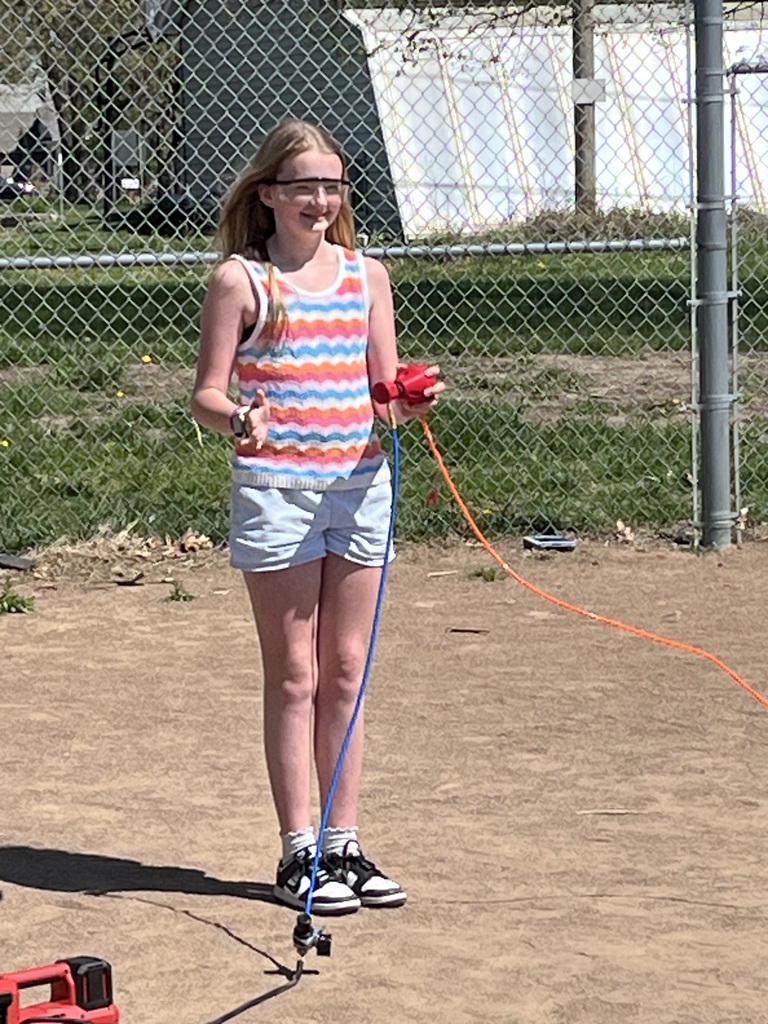 A girl in a colorful striped tank top and safety glasses smiles while holding a red rocket launcher.