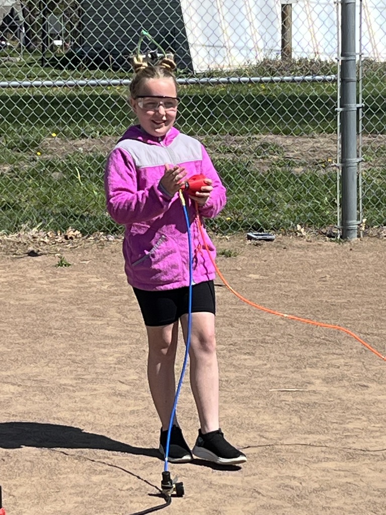 A girl in a pink fleece jacket and safety glasses smiles while preparing to press the launch button.