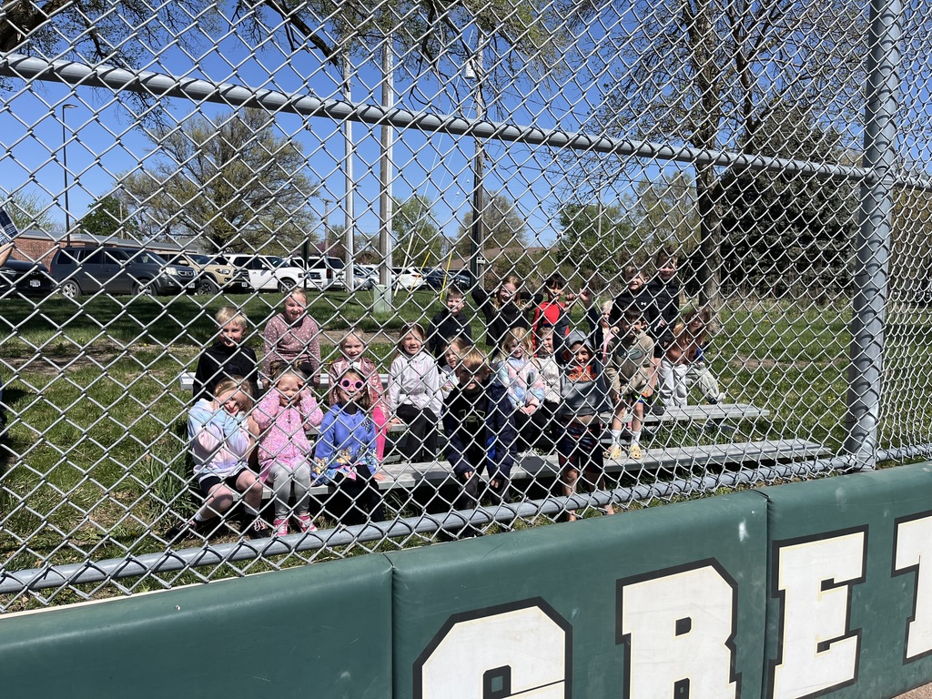 A large group of young children sit on bleachers behind a green padded fence in a park.