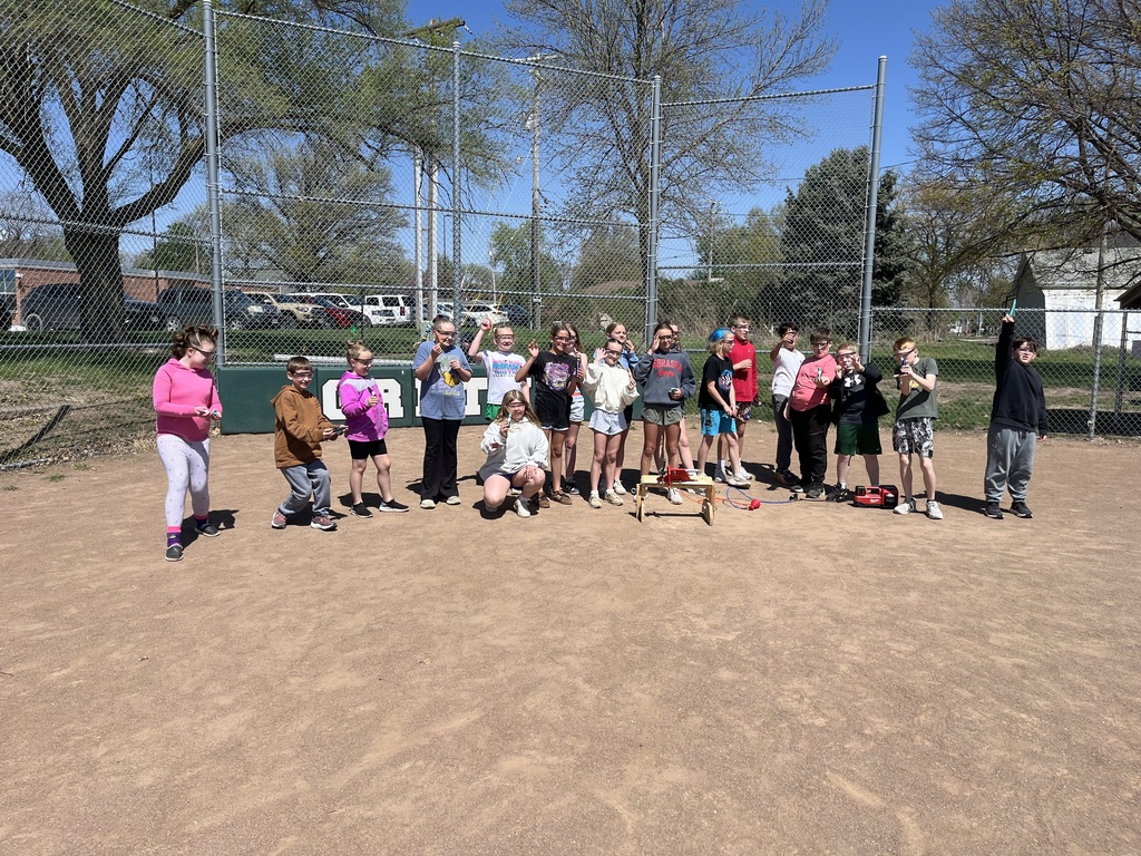 A wide group shot of students standing on a dirt field with their rockets, a red air compressor, and launch equipment.