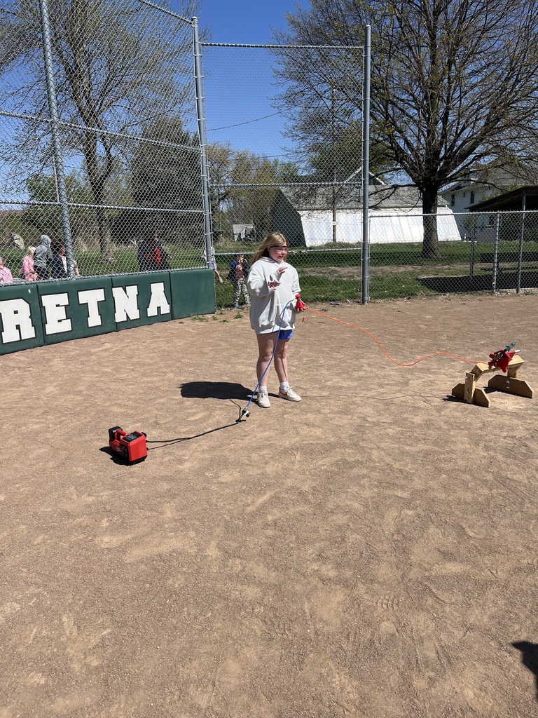 A young girl in a grey hoodie stands on a dirt field, holding a red launch controller connected to a small rocket on a wooden stand.