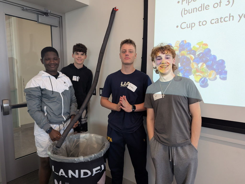 Four boys stand in front of a white board and smile for the camera. The one on the left has a piece of tubing. 