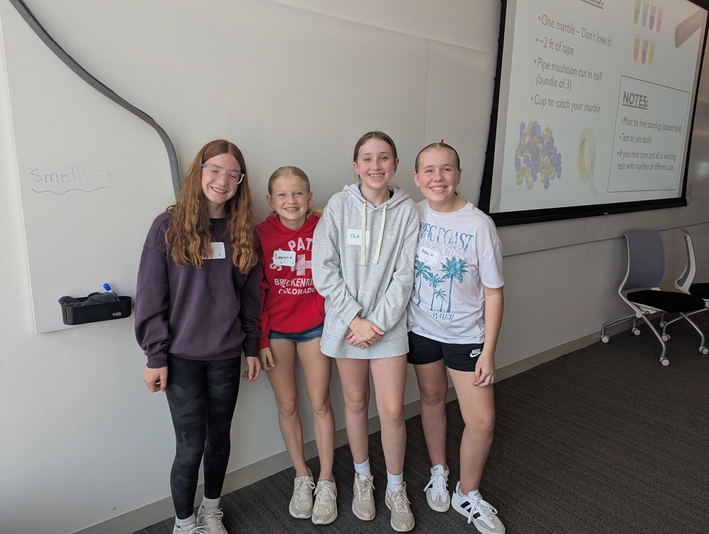 Four girls stand in front of a white board and smile at the camera. 