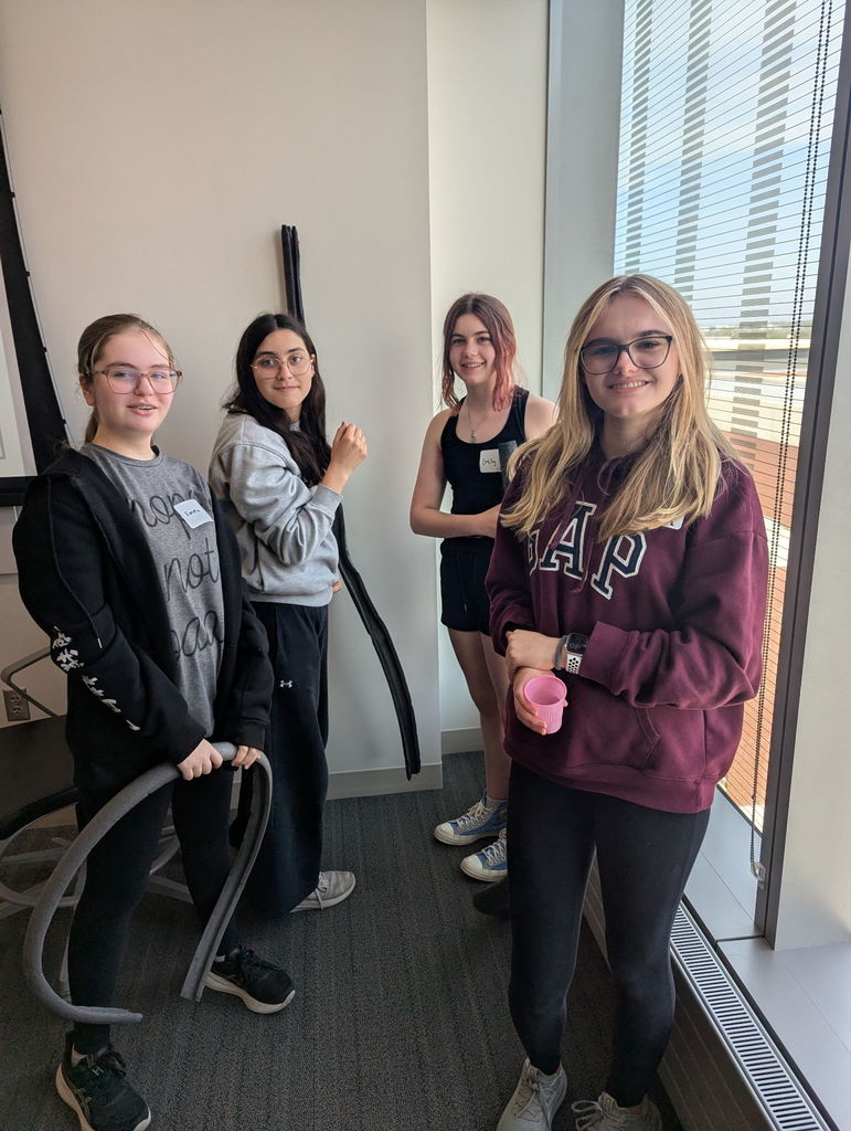 Photo of four female students standing and looking at the camera holding cups and hoses for their activity.