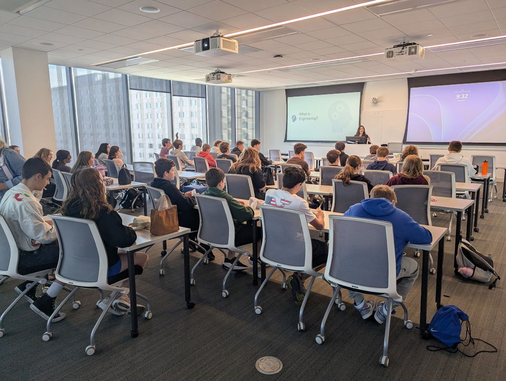 Photo taken from the back of a classroom where students are seated in chairs at tables listening to a female speaker who is presenting at the front of the room.