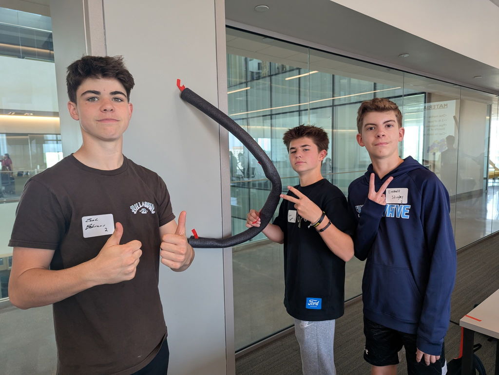Photo of three male students looking at the camera with their creation taped to the wall beside them.