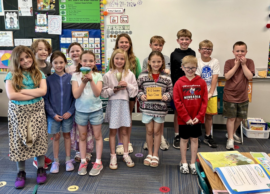  A group of eleven young elementary school students poses together in a colorful classroom. Three girls in the front row proudly display awards: a large green gemstone, a gold trophy, and a sign reading "K-2 Golden Tech Class." The background features educational posters, a calendar, and classroom decorations.