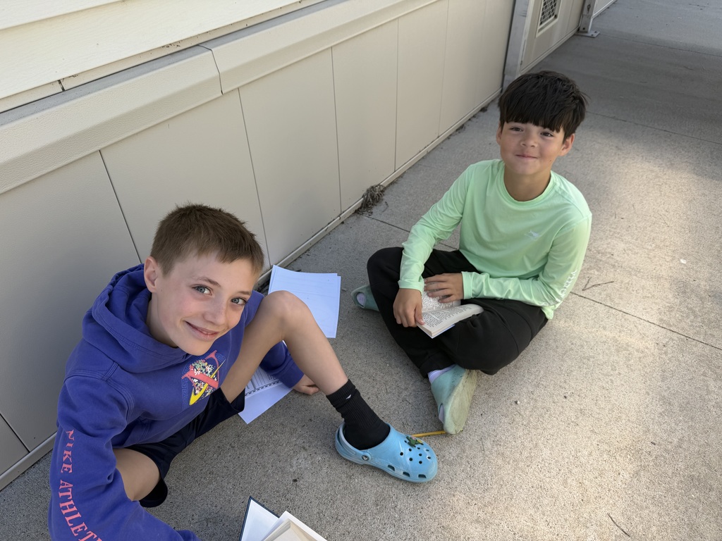 A close-up of two boys sitting on the pavement, looking up and smiling while holding their reading materials.