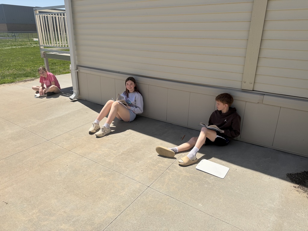 Three students sit along a shaded section of a building foundation, spread apart while reading books on a sunny day.