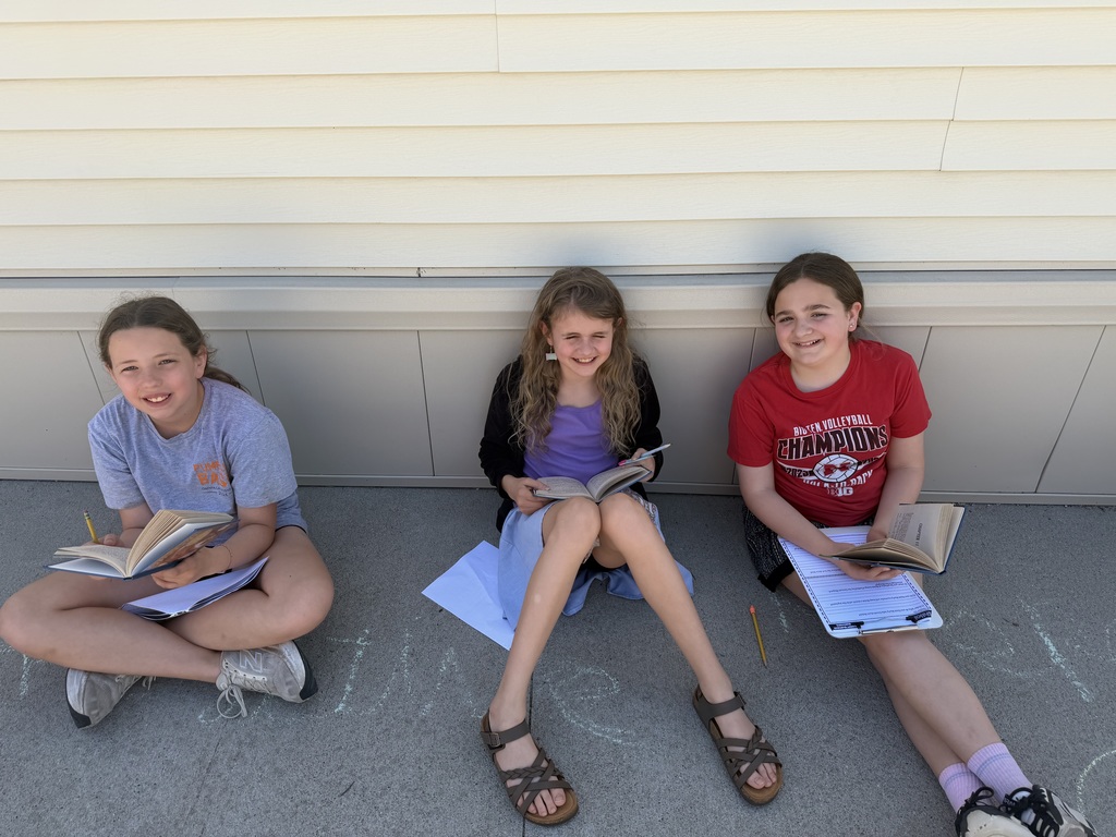 Three girls sit cross-legged against a beige building wall outdoors, smiling while holding open books and clipboards.
