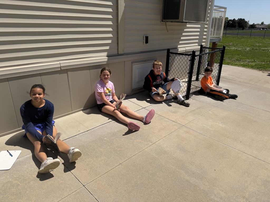 Four students are spaced out sitting against a long exterior wall in the sun, each engaged in reading or writing.
