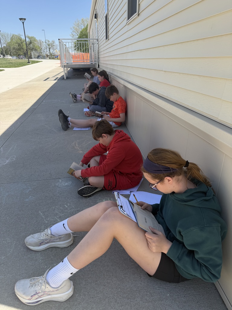 A line of several students sit with their backs against a building, focusing on reading books and writing on papers during an outdoor lesson.