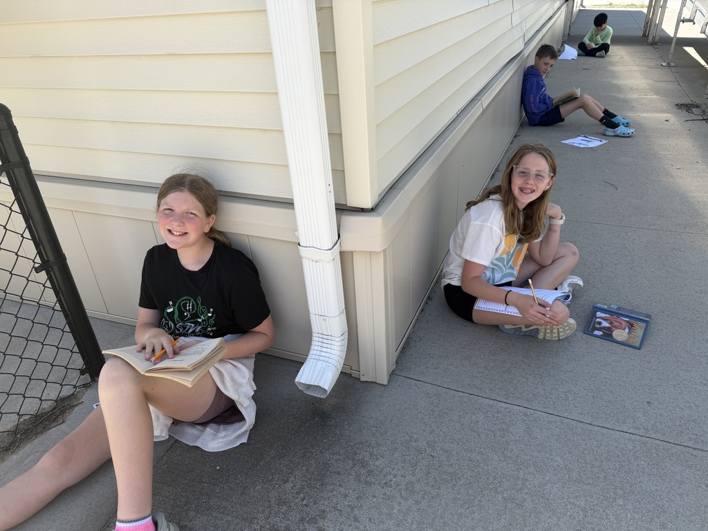 Two girls sit on the ground near a white downspout; one smiles at the camera with a book, while others are visible further down the walkway.
