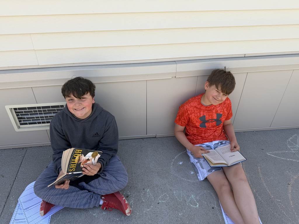 Two boys sit on a concrete walkway against a wall, both smiling and looking at the camera while holding open copies of the book Shiloh.